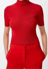 Top de punto rojo con cuello alto y ajustado y mangas cortas. Textura lisa y patrón acanalado en todo el cuerpo, combinado con pantalones rojos.