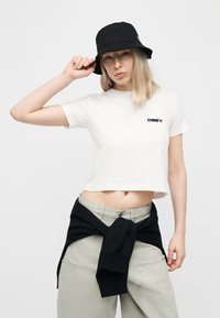 Νεαρή γυναίκα φοράει μαύρο καπέλο τύπου bucket, άσπρο κοντομάνικο μπλουζάκι με το λογότυπο "OBEY", μπεζ παντελόνι και μαύρο φούτερ δεμένο γύρω από τη μέση της.