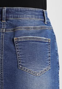 Džínová sukně s tmavě modrou barvou, která má zadní kapsu přišitou kontrastním stehem a hladkou texturu.