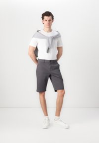 Graue Shorts aus einer Baumwollmischung mit flacher Vorderseite, Taschen und umgeschlagenen Säumen. Kombiniert mit einem weißen T-Shirt und einem grauen Pullover, der um den Hals gebunden ist.