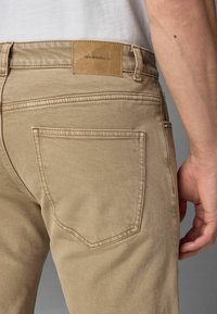 Detailný záber na osobu v béžových džínoch so zadnou vreckom a koženou nášivkou označenou "DENIM SUPPLY Co." na páse.