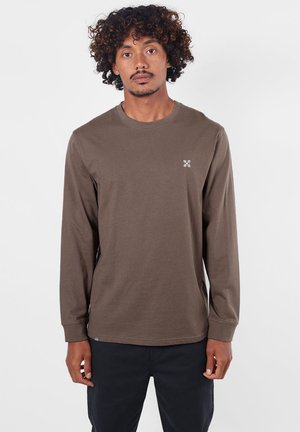 Homme aux cheveux bouclés portant une chemise marron unie à manches longues et un pantalon noir, debout sur un fond blanc uni.