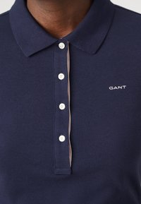 Tumed sinine polo särk tekstuurse kangaga, millel on krae, viis valget nuppu ja rinnal väike valge GANTi logo.