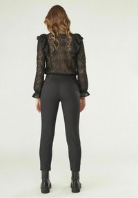 Femme avec des cheveux ondulés portant un chemisier noir en dentelle à manches longues avec des volants, un pantalon noir ajusté et des bottines noires, se tenant de dos.