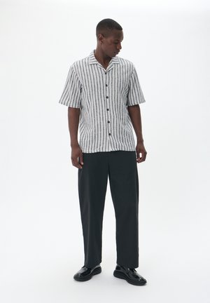 Short-sleeve shirt with vertical black and white stripes, button-down front, and a lightweight fabric. Paired with relaxed black trousers and shoes.