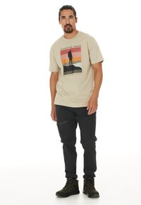 Beige grafisk t-shirt med trycket "Äventyr Framåt", tillsammans med mörka byxor. Svarta vandringskängor med snörning och dragkedjedetalj.