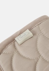 Tissu beige rembourré avec un motif matelassé en forme de coquille, comportant une petite étiquette avec un design de nuage et du texte. Texture douce avec des coutures visibles.
