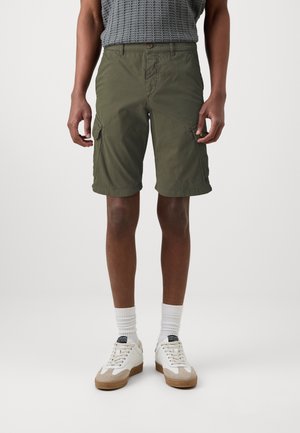 Mann trägt olivgrüne Cargo-Shorts, weiße Sportsocken und weiße Turnschuhe vor einem schlichten hellen Hintergrund.