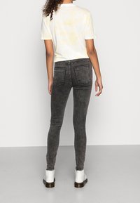 Una persona con cabello rizado lleva una camiseta de tie-dye clara y jeans negros ajustados, de espaldas, de pie sobre un fondo gris claro.