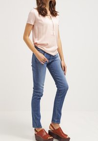 Frau trägt ein hellrosa T-Shirt mit kurzen Ärmeln, blue slim-fit Jeans und braune Plattform-Sandalen mit offenem Zeh, steht vor einem schlichten Hintergrund.
