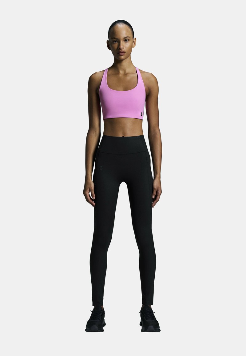 Rosa Sport-BH und schwarze Leggings mit hohem Bund. Der BH hat ein breites Tragegestell; die Leggings sind schmal geschnitten mit einer strukturierten Oberfläche. Schwarze Sportschuhe.