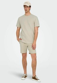 Camiseta de punto de color beige claro con mangas cortas y pantalones cortos a juego, ambos con un patrón texturizado. Combinado con una gorra beige y zapatillas con suela marrón.