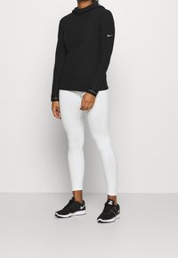 Maglietta a maniche lunghe con cappuccio nera e texture, con logo Nike dorato, abbinata a leggings bianchi aderenti e scarpe da ginnastica nere.