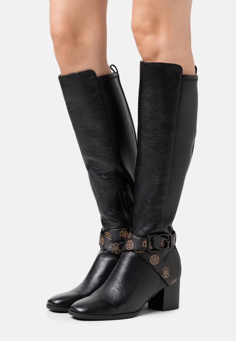 Personne portant des bottes noires en cuir jusqu’au genou avec des talons blocs et des lanières marron à logo avec boucles, debout sur fond blanc.