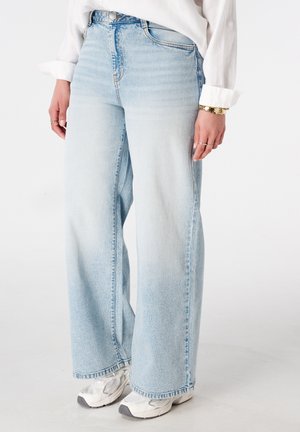 Personne portant un jean bleu clair à jambes larges, des baskets blanches et une chemise blanche à manches longues avec un bracelet en or et des bagues visibles sur les mains.