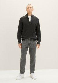 Dunkelgrauer Zipkragenpullover mit strukturierten Details, getragen über einem weißen Hemd, kombiniert mit grauen Jeans und weißen Sneakern. Das Model steht aufrecht.