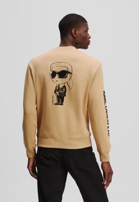 Beige pulóver, hátán fekete napszemüveges mesehős képpel, az ujjon pedig függőlegesen a "KARL LAGERFELD" felirattal.