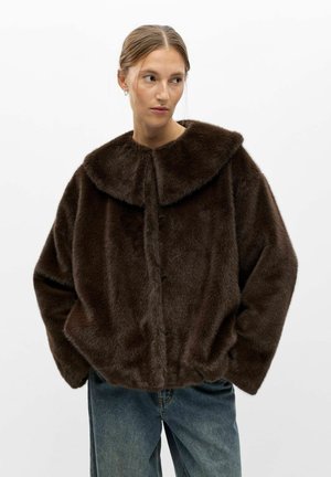 Femme à la peau claire et aux cheveux attachés en chignon bas, portant une veste en fausse fourrure marron foncé et un jean bleu, regardant vers sa droite avec les mains dans les poches.