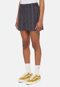 Pantalones cortos a cuadros en color navy con cinturilla elástica; tejido ligero; cuenta con líneas rojas y blancas; combinados con zapatos amarillos con cordones y calcetines blancos.