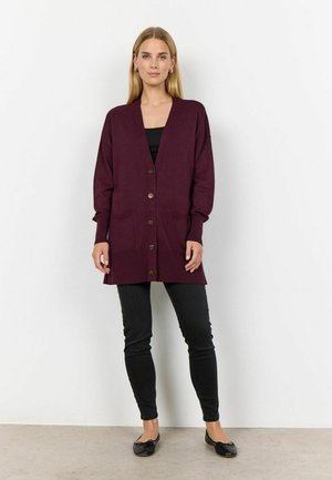 Cardigan lungo bordeaux in maglia morbida con scollo a V, bottoni frontali e due tasche. Indossato sopra una maglietta nera, abbinato a pantaloni skinny neri.