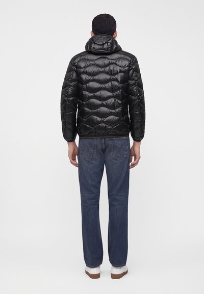 Homme de dos portant une veste matelassée noire avec capuche, un jean bleu et des baskets blanches sur un fond uni.