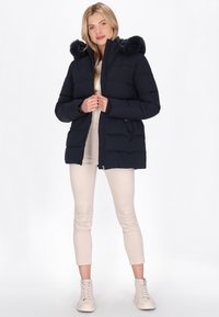Marineblauer Pufferjacke mit einer Kunstfellkapuze, ausgestattet mit einem gesteppten Design, Reißverschluss und Seitentaschen, kombiniert mit beigen, eng anliegenden Hosen und weißen Sneakers.