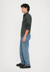 Camisa ajustada de color verde oscuro, jeans claros y zapatillas negras. La camisa tiene un corte slim, mangas largas y una textura suave.