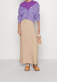 Lila stickad cardigan över en lila topp, parat med en beige ribbad maxikjol, bruna platta klackar och en liten beige handväska.