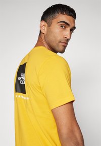 Gelbes Kurzarm-T-Shirt mit Rundhalsausschnitt. Auf der Rückseite befindet sich ein schwarzes quadratisches Logo mit weißem Text, aus weichem, leichtem Stoff gefertigt.