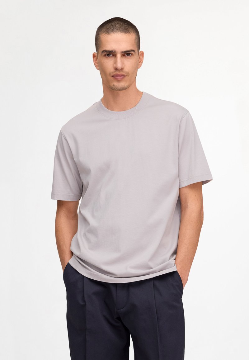 Homme aux cheveux courts portant un t-shirt gris clair à manches courtes et un pantalon bleu marine foncé, debout les mains dans les poches.