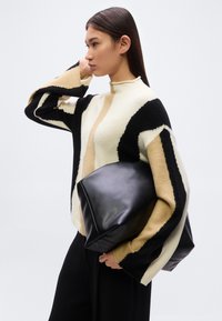 Pull en tricot crème, noir et beige avec un col montant, des manches allongées et des motifs géométriques ; assorti à un sac en cuir noir.
