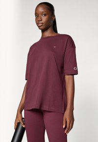 Bord rød bomullst-skjorte med kort erm, med en liten logo på brystet. Kombinert med matchende leggings, holdt med en svart vannflaske.