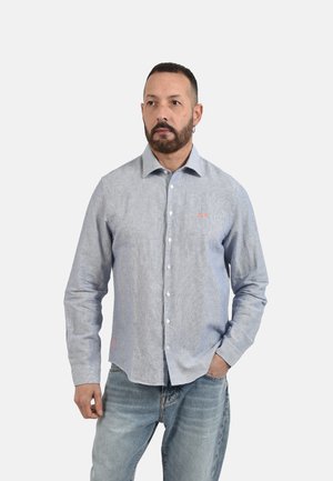 Uomo con capelli corti e barba, che indossa una camicia azzurro chiaro abbottonata e jeans blu sbiaditi, in piedi con una mano in tasca, sfondo neutro.