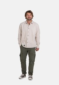 Hellbeige Button-Up-Hemd mit langen Ärmeln, kombiniert mit olivegrünen Cargo-Hosen. Die Schuhe sind braun mit weißen Akzenten und Schnürsenkeln.