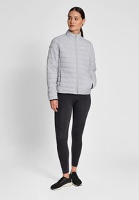 Lysegrå pudejakke med høj krave, lynlås og quiltet tekstur. Båret med sorte leggings og sneakers. Enkel branding synlig.