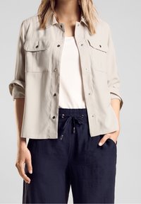 Kvinna klädd i en beige knapplång jacka med fickingar, vit topp och mörkblå dragsko-byxor, stående med en hand i fickan.