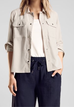 Femme portant une veste beige à boutons avec des poches à rabat, un haut blanc et un pantalon bleu marine à cordon, debout avec une main dans la poche.