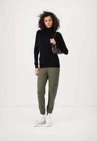 Jersey de cuello alto negro, pantalones jogger verde oliva con bolsillos con cremallera y zapatillas blancas. El cabello rizado y un pequeño bolso marrón completan el look.