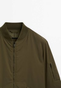 Veste bomber vert olive avec col côtelé, fermeture éclair à l'avant et poche zippée sur la partie supérieure de la manche gauche, présentée sur fond blanc.