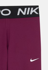 Fioletowe szorty sportowe z czarnym pasem, na którym widnieje częściowy biały napis "NIKE" oraz biały logo swooshu Nike na nodze.