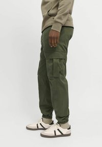 Person iført olivengrønne cargobukser med elastiske manchetter og beige sneakers med sorte striber, stående mod en hvid baggrund.