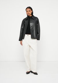 Sort jakke i kunstlæder med spidst revers og knapper foran, kombineret med hvide højtaljede bukser og sorte flade sko.