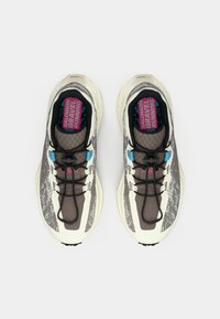 Αθλητικά παπούτσια με υφή καφέ και κρεμ άνω μέρος, μαύρα κορδόνια και πολύχρωμο πάτο με την ένδειξη "GRAVEL RUNNING".