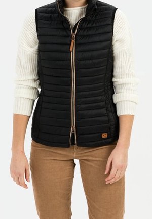 Femme portant un gilet matelassé noir par-dessus un pull en tricot blanc et un pantalon marron clair, debout devant un fond uni.