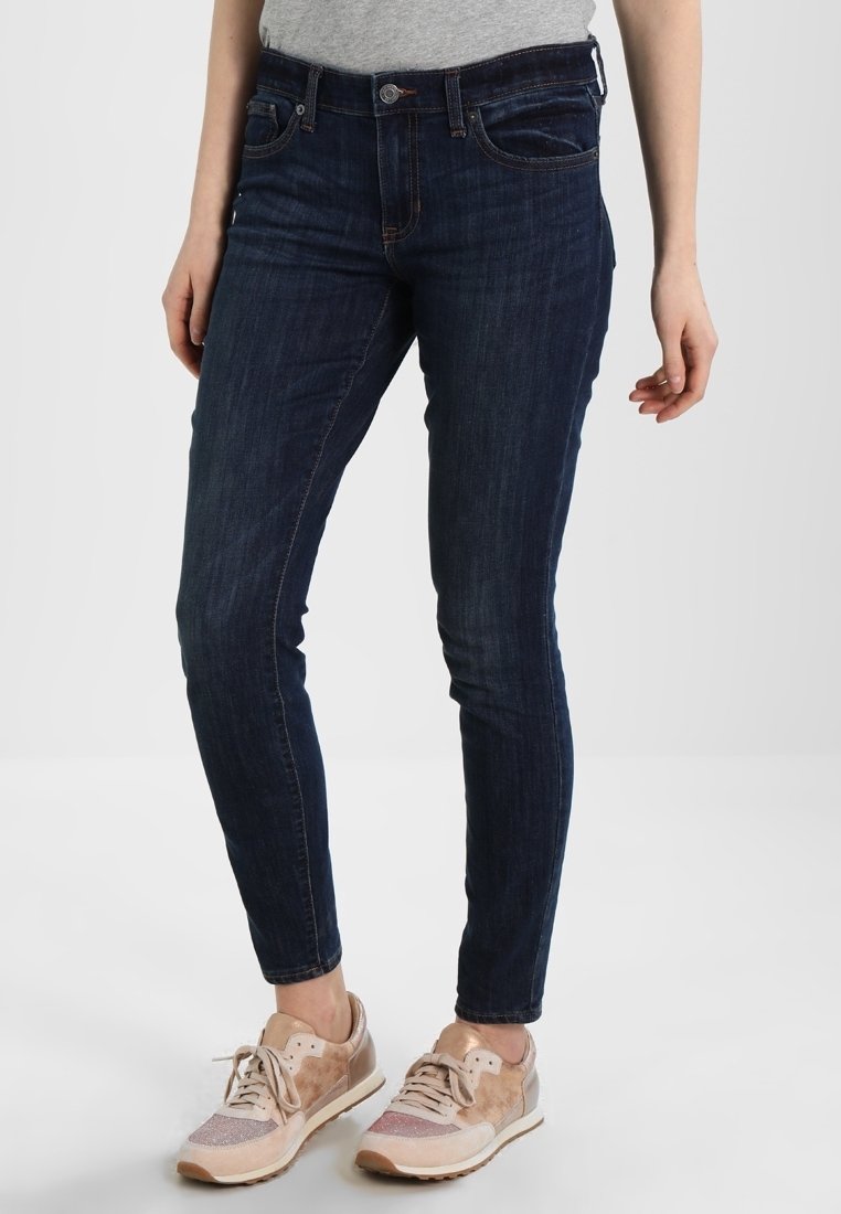 Jean slim bleu foncé porté avec des baskets beige et rose, porté par une personne debout devant un fond uni.