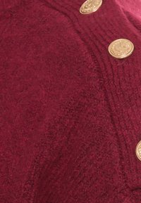 Burgundy stickad cardigan med texturerat tyg, framträdande vertikal ribbning och dekorativa knappar i guldton.