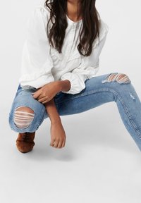 Blusa blanca con detalles de encaje, jeans rasgados de un azul claro y zapatos de ante marrones. Los jeans presentan grandes rasgaduras en las rodillas con detalles horizontales.