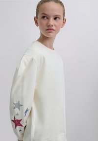 Kermanvärinen collegepaita, jossa pyöreä kaula-aukko, pitkät hihat ja tähdelliset kuviot punaisina, hopeanvärisinä ja sinisinä yksityiskohtina yhdellä hihalla.