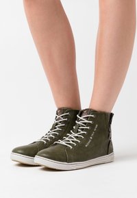 Olivgrüne hohe Sneakers aus strukturiertem Leder, mit weißen Schnürsenkeln, einem seitlichen Reißverschluss und dem Druck "MUSTANG TRUE DENIM".