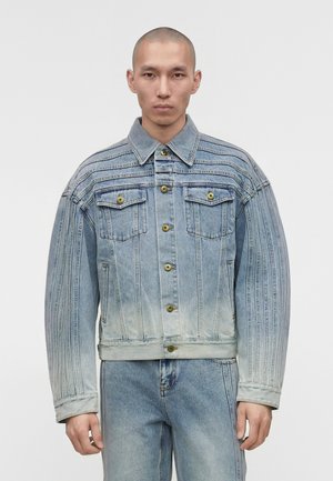 Άνδρας με ξυρισμένο κεφάλι φοράει ανοιχτόγαλαν τζιν μπουφάν με κάθετες ραφές και ταιριαστό τζιν παντελόνι σε λευκό φόντο.